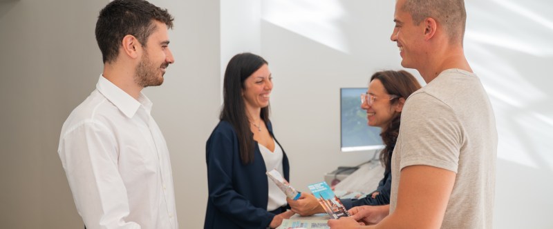 Two people at a reception desk talking to two staff members, holding brochures.