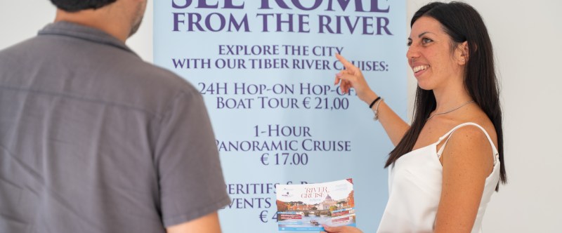 Woman points to a cruise poster while holding brochures, speaking with a man.
