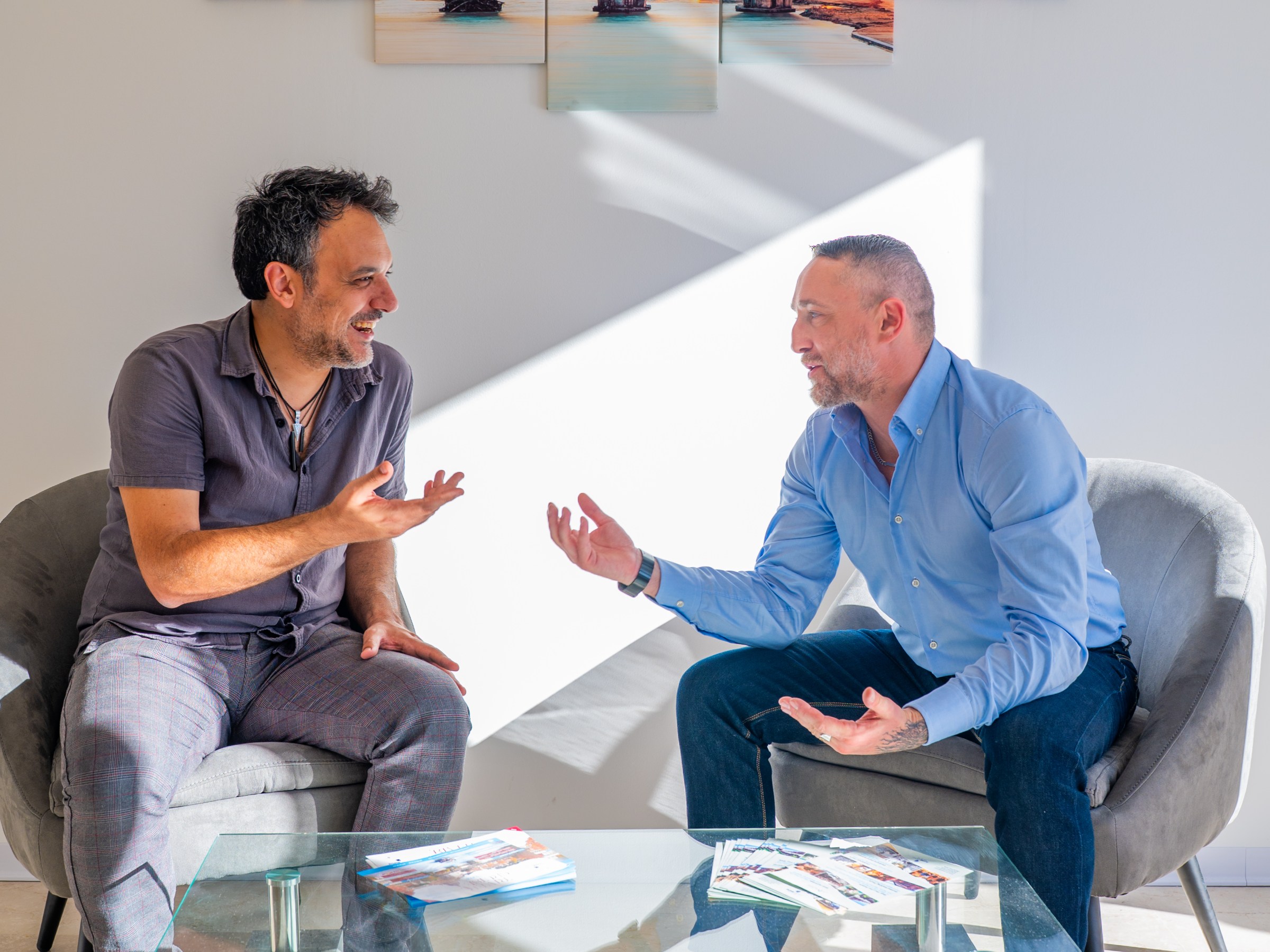Two men sitting and talking in front of a multi-panel cityscape artwork on a wall.