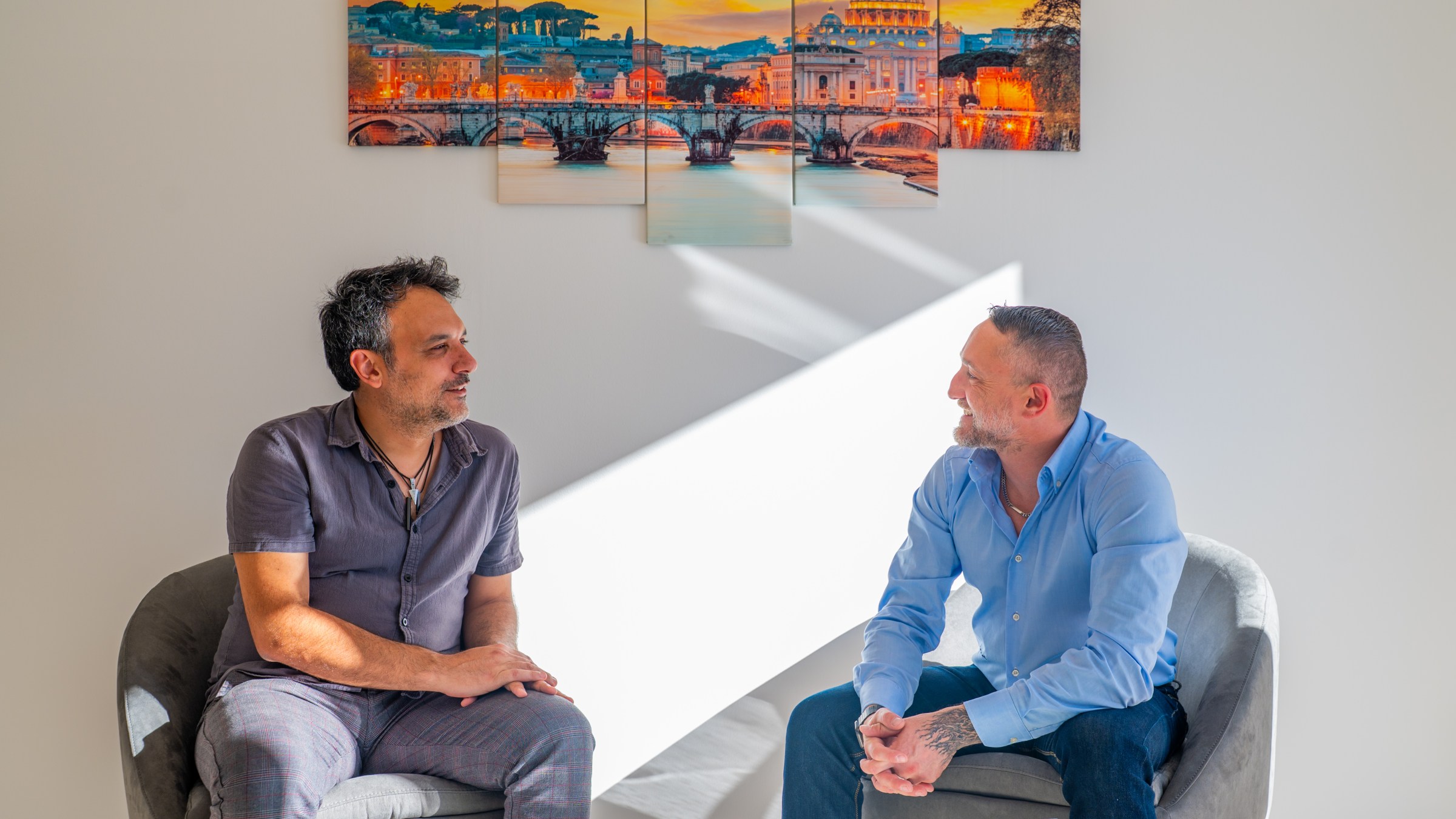 Two men sitting and talking in a room with a colorful cityscape painting on the wall.