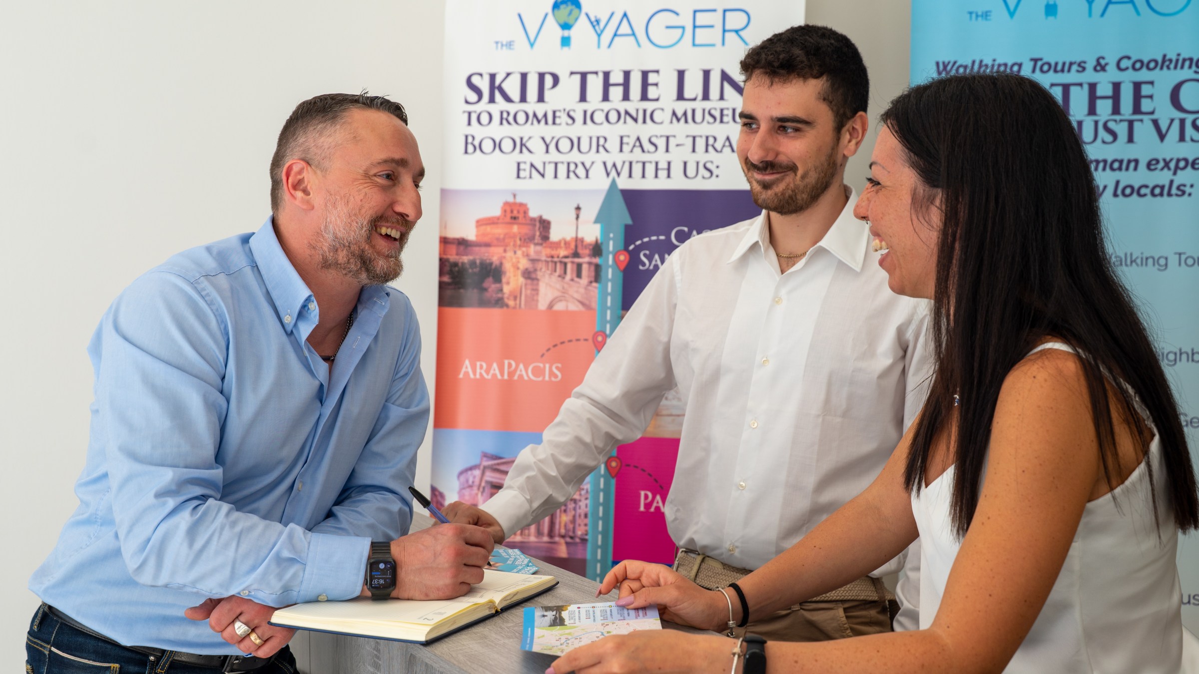 Three people smiling and talking at a travel agency counter with 'Skip the Line' posters behind them.